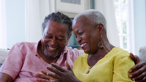 Una-Pareja-De-Ancianos-Sonriente-Sentada-Y-Hablando-En-El-Sofá-De-Casa