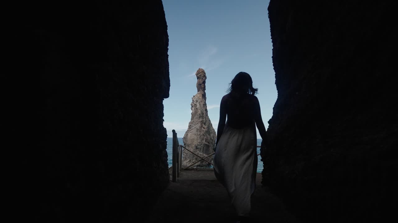 Woman in flowing dress walks toward Ilhéus rock through a natural stone corridor, framed against Madeira’s blue ocean and bright afternoon sky.