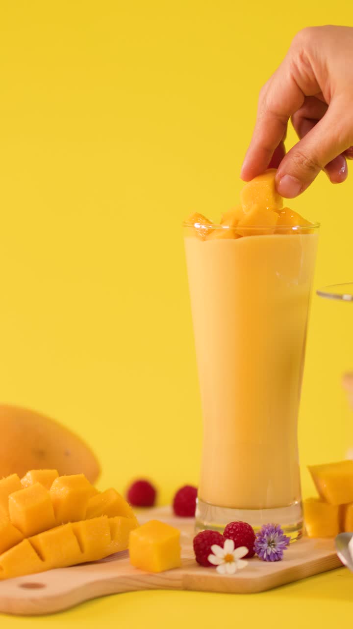 Primo piano di una mano che aggiunge cubetti di mango a un alto bicchiere da frullato su uno sfondo giallo vivace.