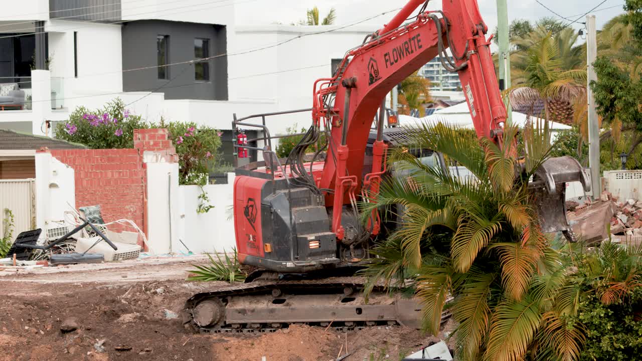 A red excavator removes palm plants and soil at a residential construction site in daylight, with steady camera and modern Gold Coast homes in background