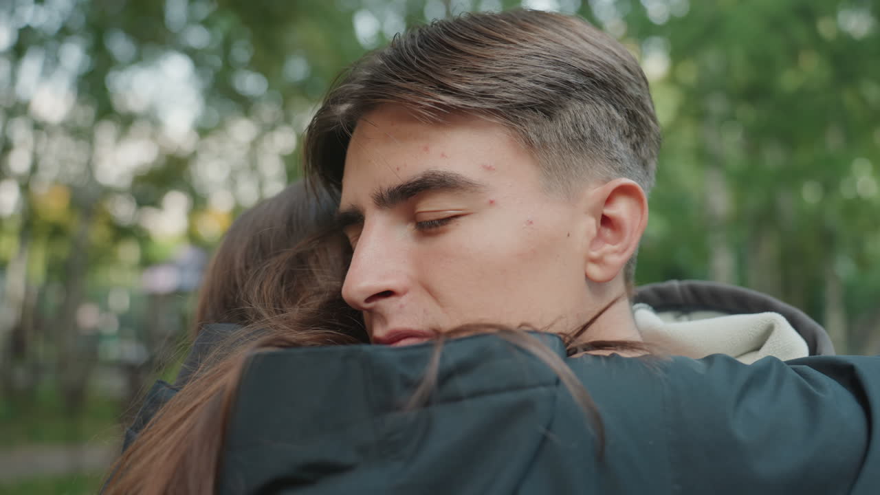 Giovane coppia caucasica si abbraccia in un parco sotto alberi verdi, con zaini e giacche visibili, luce pomeridiana soffusa, occhi chiusi in un abbraccio confortante, tranquillo sentiero, sincero ricongiungimento ed emozione
