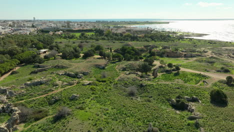 the archaeological splendor of the tombs of the kings,expansive green fields and ancient ruins leading towards the modern cityscape of paphos, juxtaposed against the clear blue waters of the sea