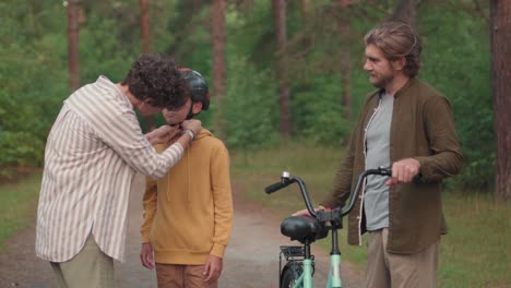 La-Madre-Le-Pone-El-Casco-A-Su-Hijo-Mientras-Su-Padre-Sostiene-Su-Bicicleta,-Luego-El-Niño-Monta-Su-Bicicleta-En-El-Bosque