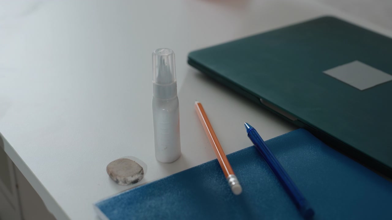 Primo piano scrivania, quaderno, flacone spray, mano che afferra gomma e matita, cartella verde su tavolo con tappetino blu, spazio di lavoro ordinato per tutor domestico, preparazione esercizi di grammatica, luce naturale soffusa, calma, concentrata.