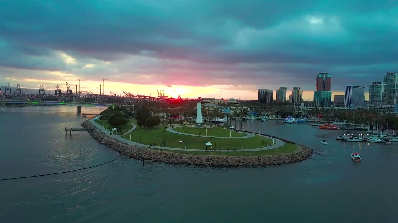 Aerial pull back of Light house in the Long Beach Harbor showing downtown buildings, at sunset with dark clouds