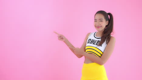 분홍색 배경에 밝은 노란색 치어리더 복장을 한 아시아 여성은 왼쪽의 빈 카피 공간을 향해 양손을 들고 있습니다.