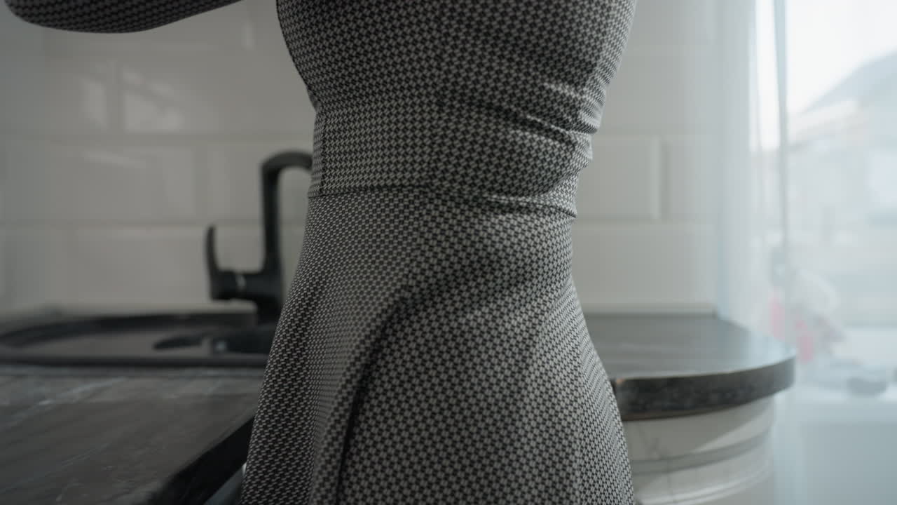 Partial view of woman wearing plaid gown in bright kitchen setting carefully placing glass cup on countertop near sink with sunlight streaming through sheer curtain