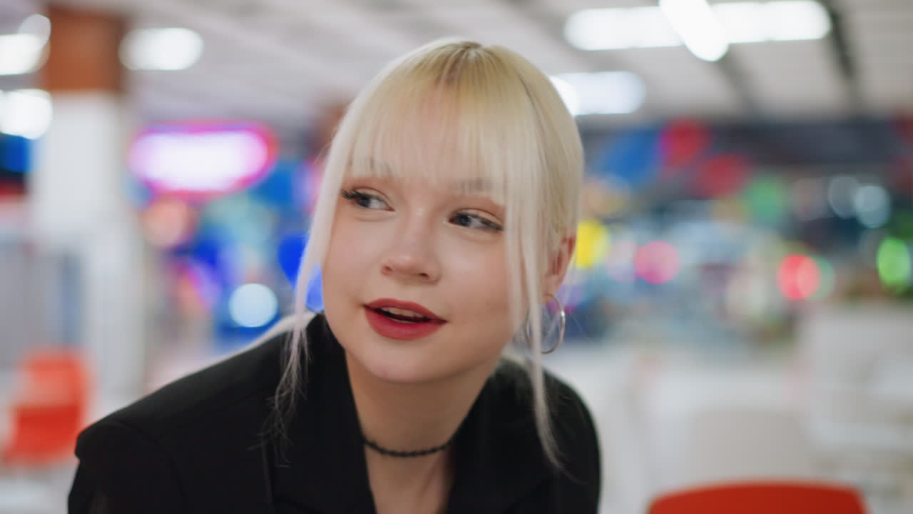 Close up of young blonde woman with painted lips smirking gently while looking around, wearing hoop earrings and black outfit, showing elegance, casual lifestyle, and beauty expression