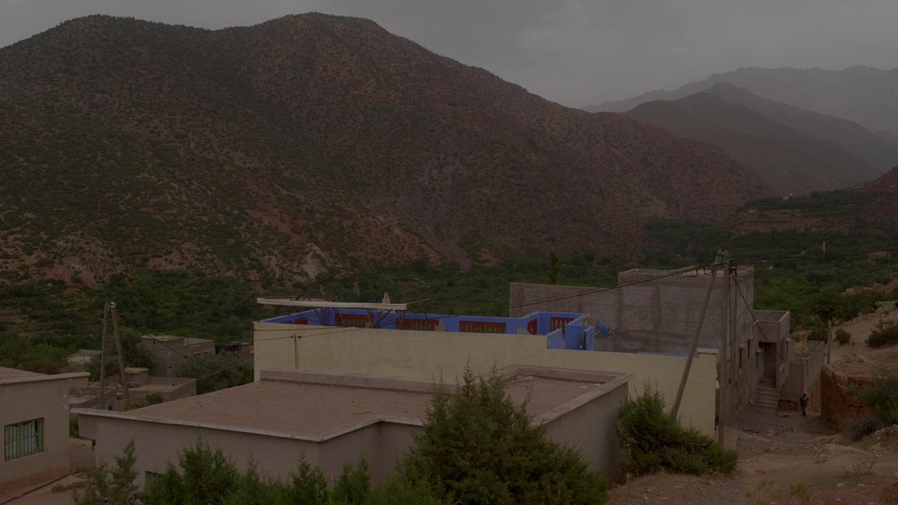Una casa di villaggio tradizionale presenta vivaci pareti sul tetto dipinte di blu sullo sfondo di spettacolari vette montuose aride e valli verdeggianti nella remota regione dell'Alto Atlante in Marocco