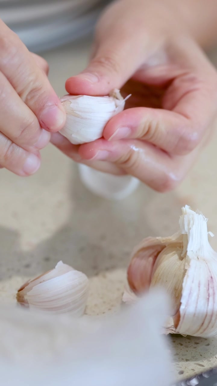 Primo piano di mani che sbucciano spicchi d'aglio su un piano di lavoro della cucina con illuminazione naturale e un'atmosfera tranquilla.