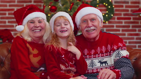 Senior-grandparents-with-granddaughter-in-Santa-hats-laughing-out-loud-at-home-near-Christmas-tree