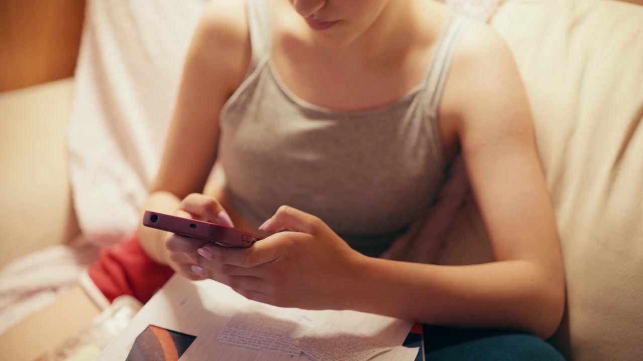 Ragazza delle scuole superiori concentrata, seduta su un divano, digita sul suo telefono cellulare, usando uno smartphone per copiare appunti, cercare risposte d'esame o barare a un test futuro mentre studia a casa.