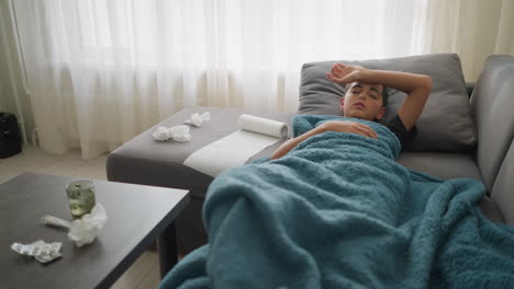 sick child resting on couch with blanket, covering mouth while coughing, used tissues on table with thermometer and glass cup, adjusting for comfort in living room during illness