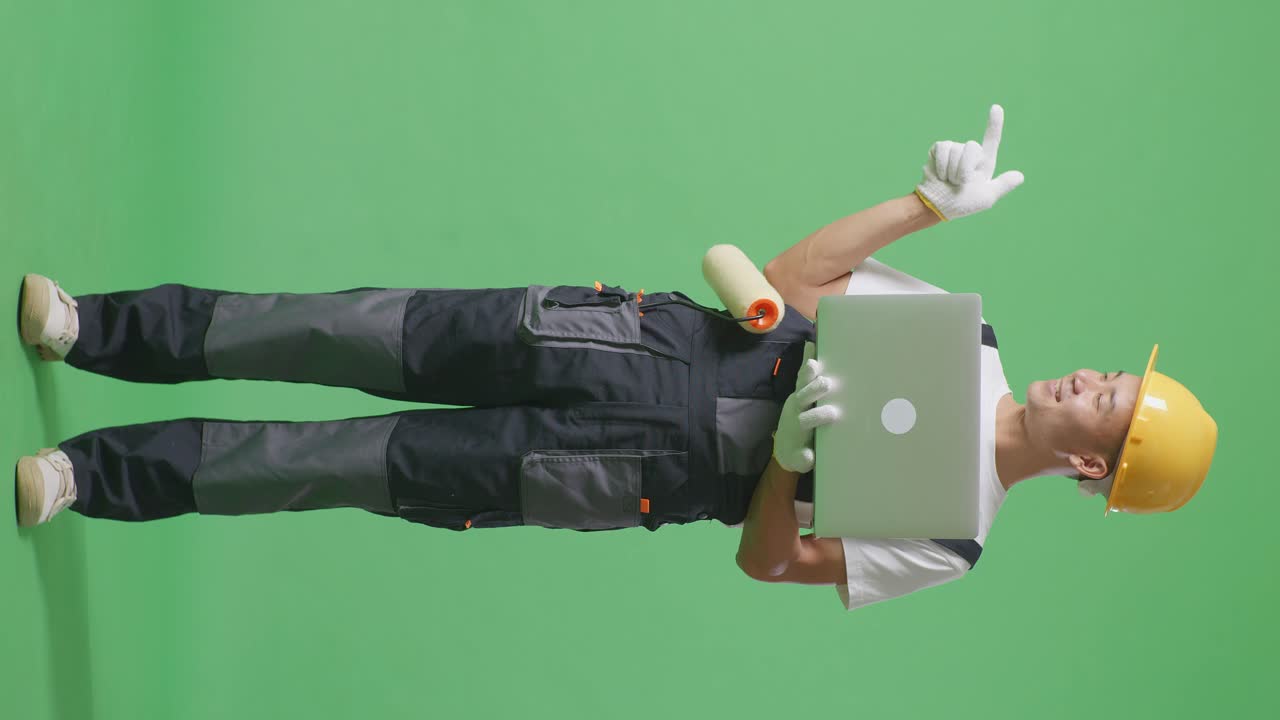 Full Body Of Asian Man Painter Wearing Safety Helmet Using A Laptop And Pointing To Side While Standing In The Green Screen Background Studio