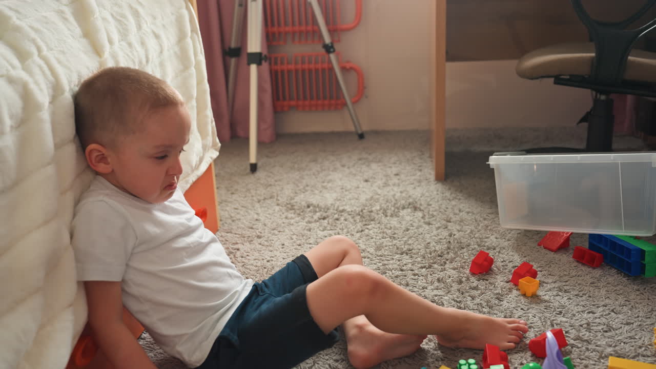 Bambino circondato da giocattoli. Il bambino riflette tranquillamente tra giocattoli d'infanzia sparsi e luce. Il figlio siede pensieroso accanto a blocchi giocattolo disorganizzati sotto la luce soffusa del sole pomeridiano dopo aver giocato