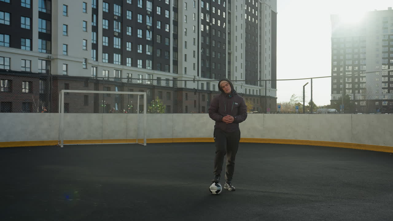hombre de pie en una arena deportiva con una pierna apoyada en una pelota de fútbol, manos juntas, cabeza ligeramente girada, rodeado de edificios urbanos bajo la brillante luz del sol