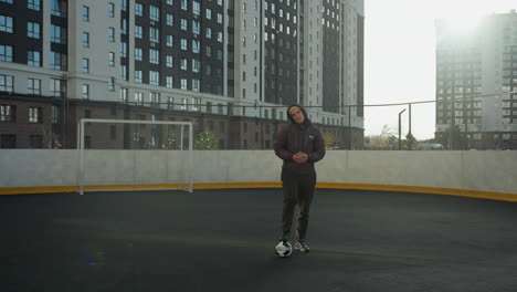 man standing on sports arena with one leg resting on soccer ball, hands clasped together, head turned slightly, surrounded by urban buildings under bright sunlight