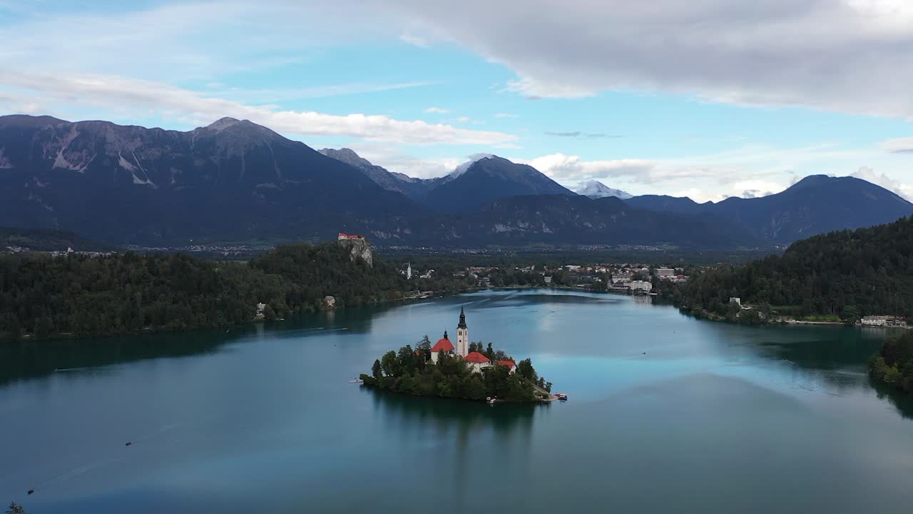A drone circles Lake Bled’s enchanting island church, capturing sweeping alpine views, shimmering blue water, and Slovenia’s breathtaking mountains rising boldly in the distance