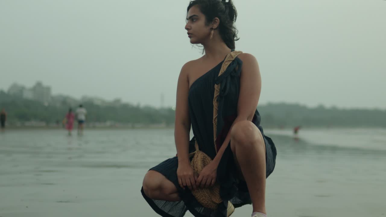 женщина в сари приседает на мокром пляже, смотрит в сторону с ветром в волосах, облачная погода