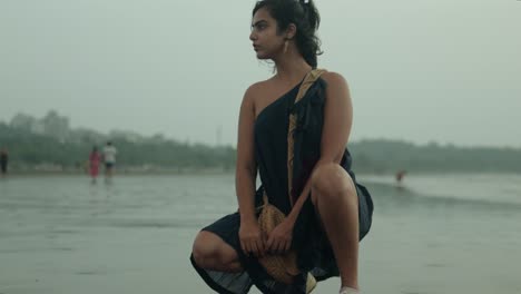 mujer con un sari en cuclillas en una playa húmeda, mirando hacia el lado con el viento en el cabello, tiempo nublado