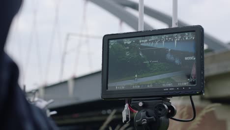 handheld camera view of a monitor on tripod showing live feed from a drone of white curved-arch bridge under overcast sky, blurred person in foreground