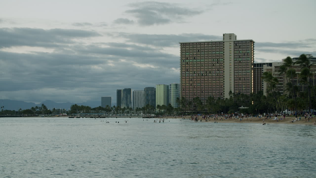 People enjoying a beach with city buildings and palm trees under a cloudy sky