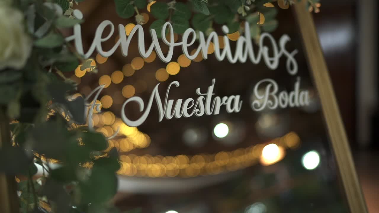 Lenta rivelazione di un cartello con la scritta “Bienvenidos a Nuestra Boda” incorniciato da fiori bianchi e vegetazione, con una calda illuminazione ambientale che crea un'accoglienza romantica, con luce LED sullo sfondo con effetto bokeh