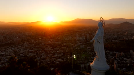 virgin mary statue on cerro san cristobal hill, overlooking santiago cityscape at golden hou, sun behind andes mountains, chile. aerial forward