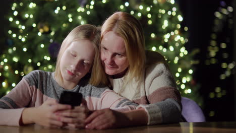 mutter und tochter schauen zusammen auf den bildschirm des smartphones. sie sitzen vor den verschwommenen lichtern des weihnachtsbaums