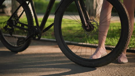 close-up of someone's legs as they kick the bicycle tire to check air pressure, the background features lush greenery, adding a vibrant touch to the scene