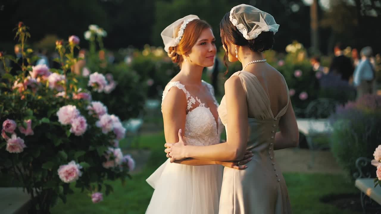 Meeting two brides in white lace, pale satin gowns embracing in rose garden, wedding portrait