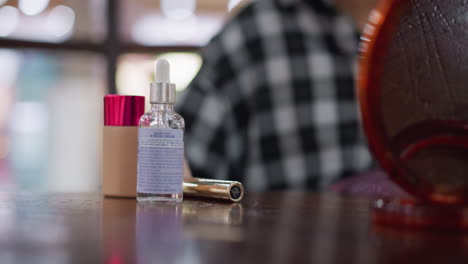 vista parcial de una mujer con camisa a cuadros arreglando herramientas de maquillaje en una mesa de madera, botella de suero de vidrio con gotero en enfoque agudo, fondo borroso cálido y productos de belleza