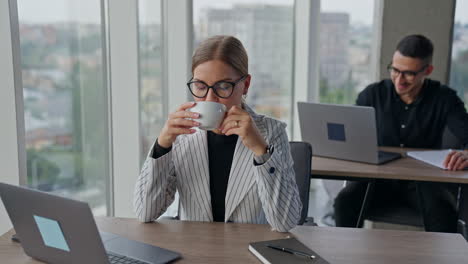 relaxed business lady enjoying coffee during break from work. lunch time at the workplace. man working at the desk behind in blurred backdrop.