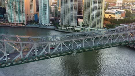 Aerial-birds-eye-view,-flyover-Brisbane-river-along-Story-bridge-capturing-vehicle-traffics-travelling-between-Fortitude-Valley-and-Kangaroo-points-with-riverside-downtown-cityscape-views-at-sunset