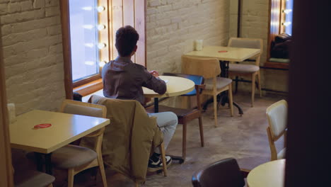 young man sitting alone at round table near window in empty cafe, coat draped over chair, back view, daytime soft light, waiting, minimal interior with chairs and backpack on bench, quiet mood