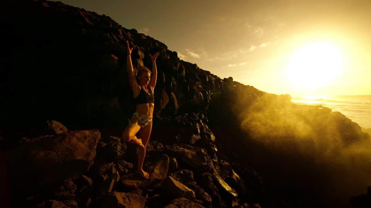 Woman practicing yoga at sunrise on volcanic rocks by the ocean