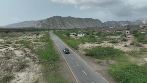 Aerial-drone-shot-over-a-car-driving-beside-village-with-mountain-range-in-the-background-in-Hingol,-balochistan,-Pakistan-on-a-cloudy-day