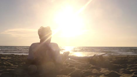 young man surfer in wetsuit lying down with surf board on the sand at the beach listening to music dancing looking at the ocean at sunset