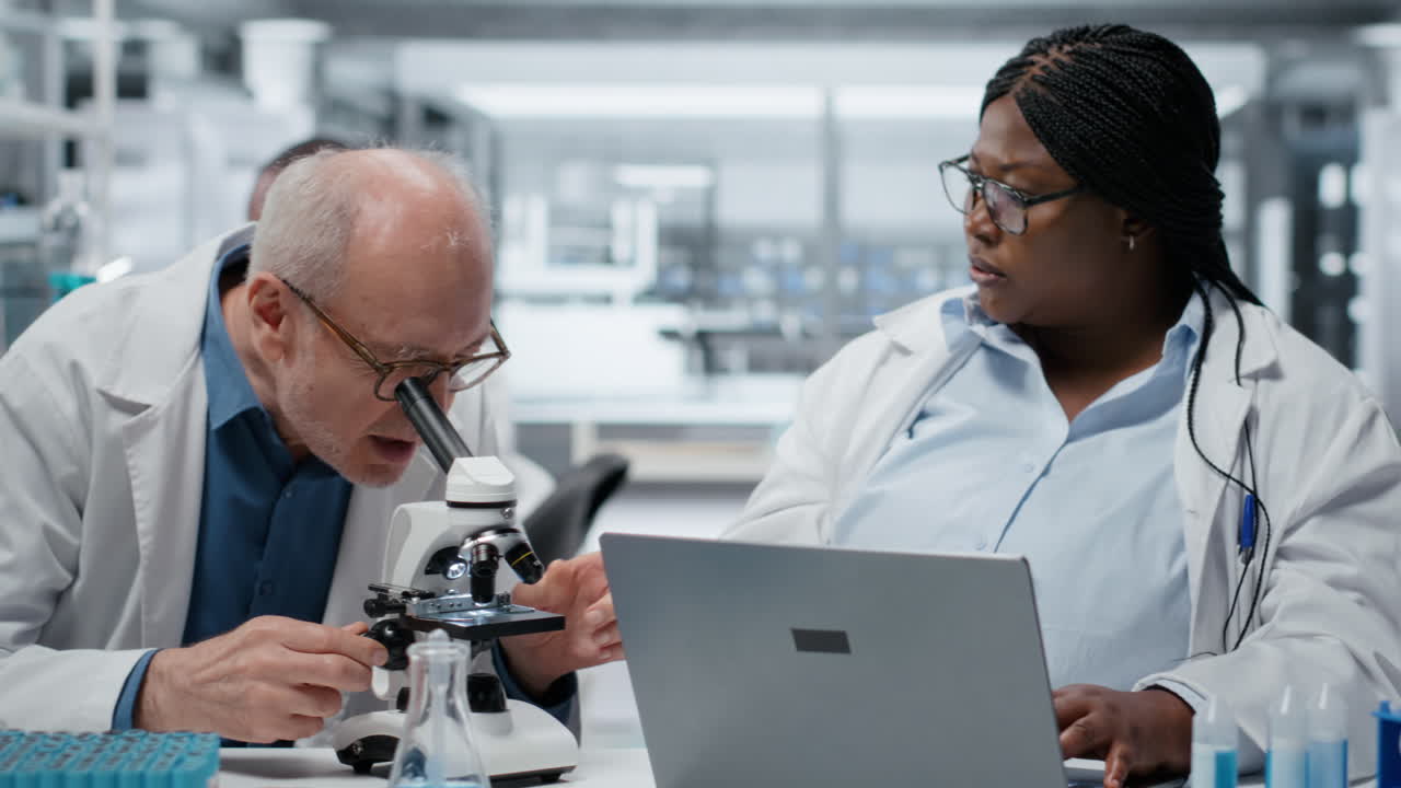 Pathologist examining biopsy slide while coworker documents findings on laptop