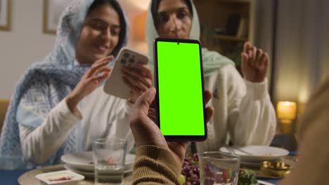 Man-Looking-At-Green-Screen-Mobile-Phone-Sitting-Around-Muslim-Family-Table-At-Home-Eating-Iftar-Meal-Breaking-Daily-Fast-During-Ramadan