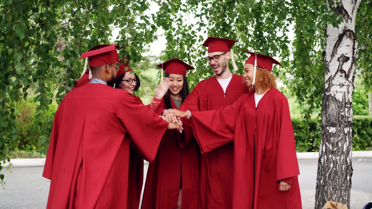 Joyful graduating students multiracial group are putting hands together then clapping hands celebrating graduation, friends are standing outdoors wearing gowns and mortar-boards.