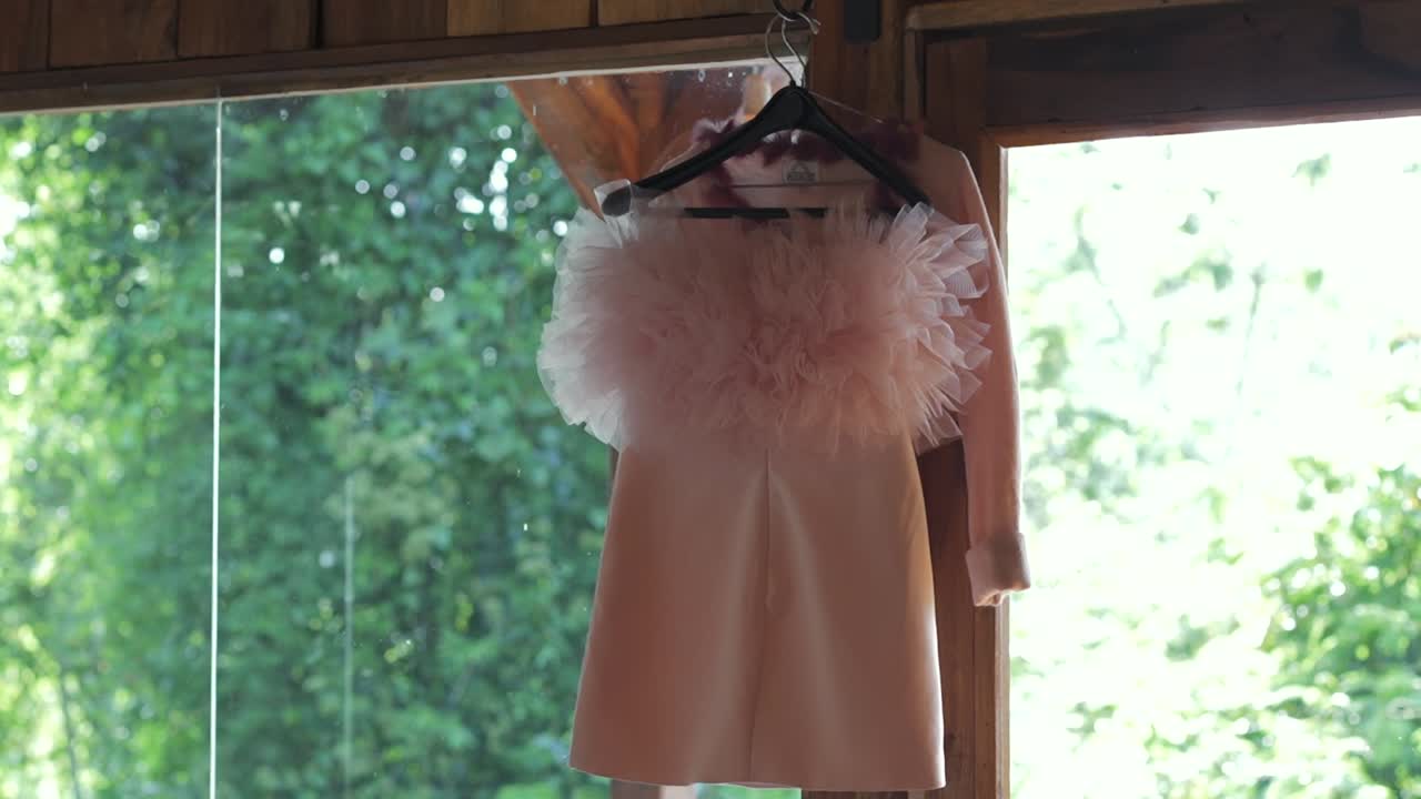 A chic, short pink wedding dress or bridal party dress with a feathered collar hangs in front of a large window with a lush green outdoor background
