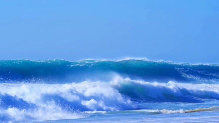 Tall turquoise swell curls and collapses into turbulent whitewater beside shallow shore, creating spray, foam, and strong coastal texture under bright daylight with open horizon, with ocean, wave
