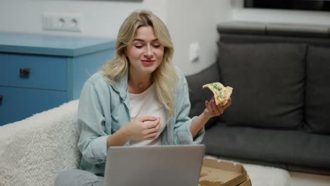 chica sentada en el sofá en la sala de estar moderna y tomando una pizza sabrosa mientras usa una computadora portátil