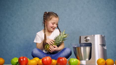 Little girl with fresh fruits. Cute child sitting on the table and playing with pineapple. Organic fruits and a squeezer on table.