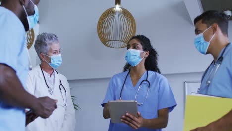 Diverse-group-of-male-and-female-doctors-in-face-masks-using-tablet-and-discussing-in-hospital