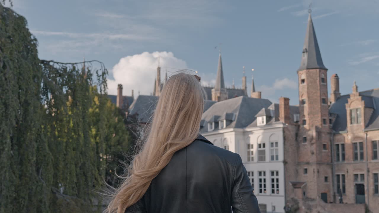 A beautiful blonde woman stands with her back to the viewer, gazing out over the tranquil canal at the iconic Rozenhoedkaai (Rosary Quay) in Bruges, Belgium