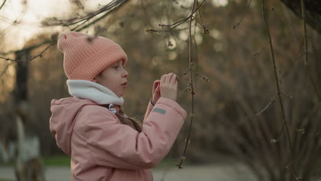 a little girl dressed in a pink jacket and beanie is seen playing with a dry tree branch, exploring her natural surroundings with curiosity. the image captures a moment of innocent childhood wonder