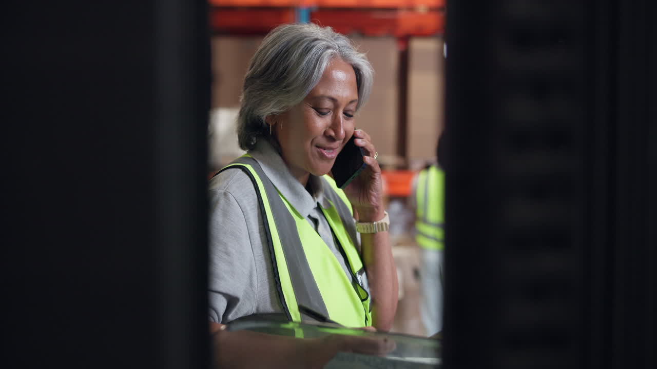 Smiling Woman in Safety Vest Talking on Phone and Writing in Warehouse