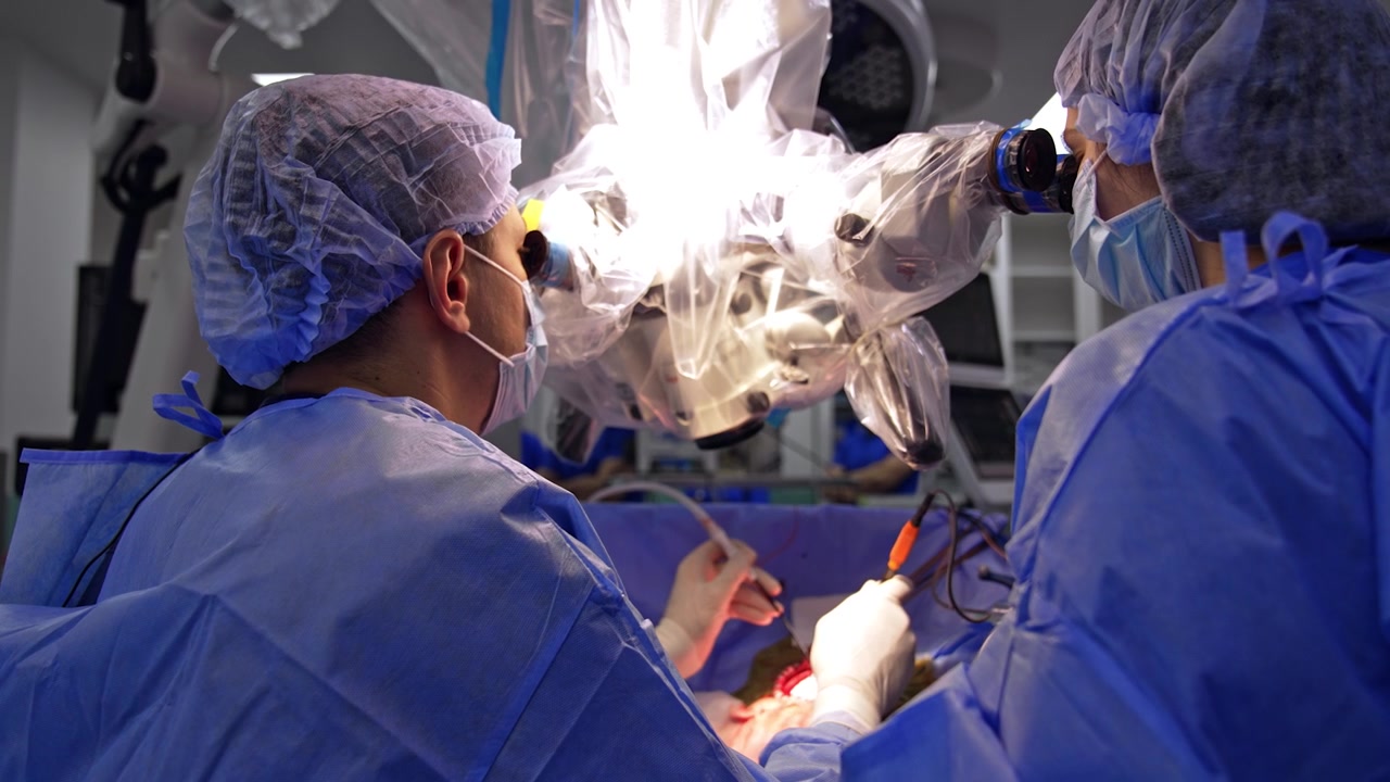 Rear view of the doctors looking at microscope performing operation. Surgeon uses tool devices and assistant sprays solution from a syringe.